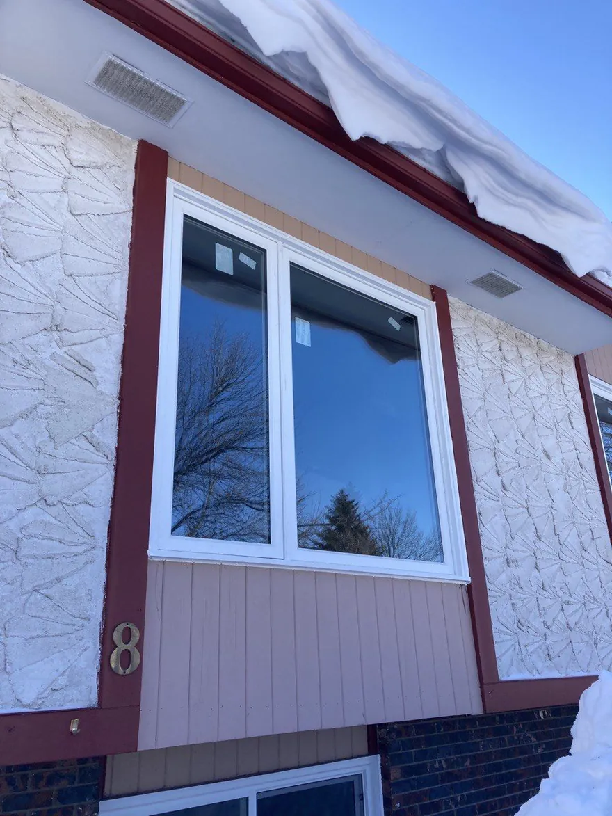 Snow hanging over the roof edge above a new casement window on a stucco house